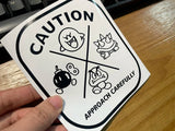 Sticker with cartoon characters and 'Caution' text held by a hand on a wooden surface.