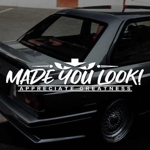 Black car with a decal that reads 'MADE YOU LOOK! APPRECIATE GREATNESS' on a blurred background.