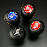 Four black gear shift knobs with different colored logos on a dark background