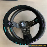 Black steering wheel with decorative elements on a light wooden surface, featuring the TopJDMStore.com logo.